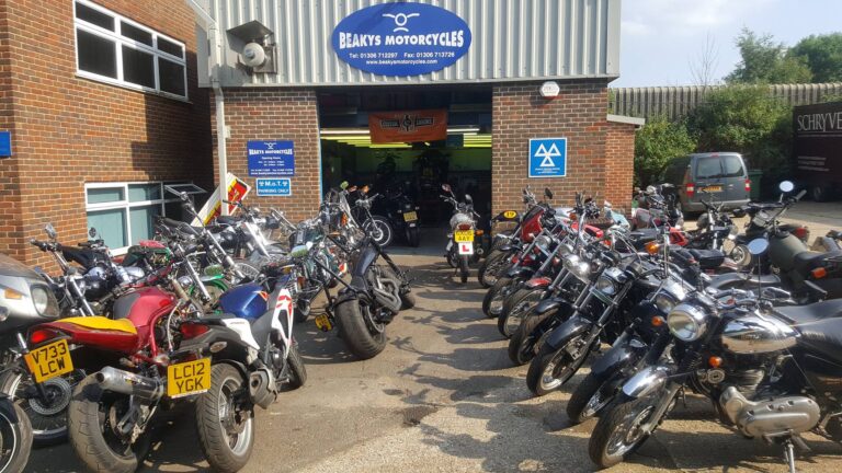 The entrance to Beaky's Motorcycle Shop under a bright sunny sky, with their logo prominently displayed above the access door. Motorcycles line each side of the driveway leading to the entrance, creating a welcoming display of two-wheeled craftsmanship